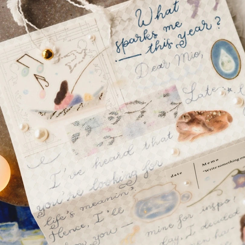 A close-up of a handmade greeting card or note with handwritten lettering, decorative washi tape, pearls, small gold beads, and natural stone accents on a neutral textured background.