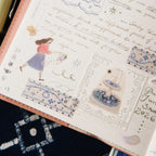A close-up of a decorative planner or journal page with pastel blue and pink illustrated stickers, handwritten script, and patterned washi tape on a peach-colored notebook cover, resting on a dark patterned fabric surface.