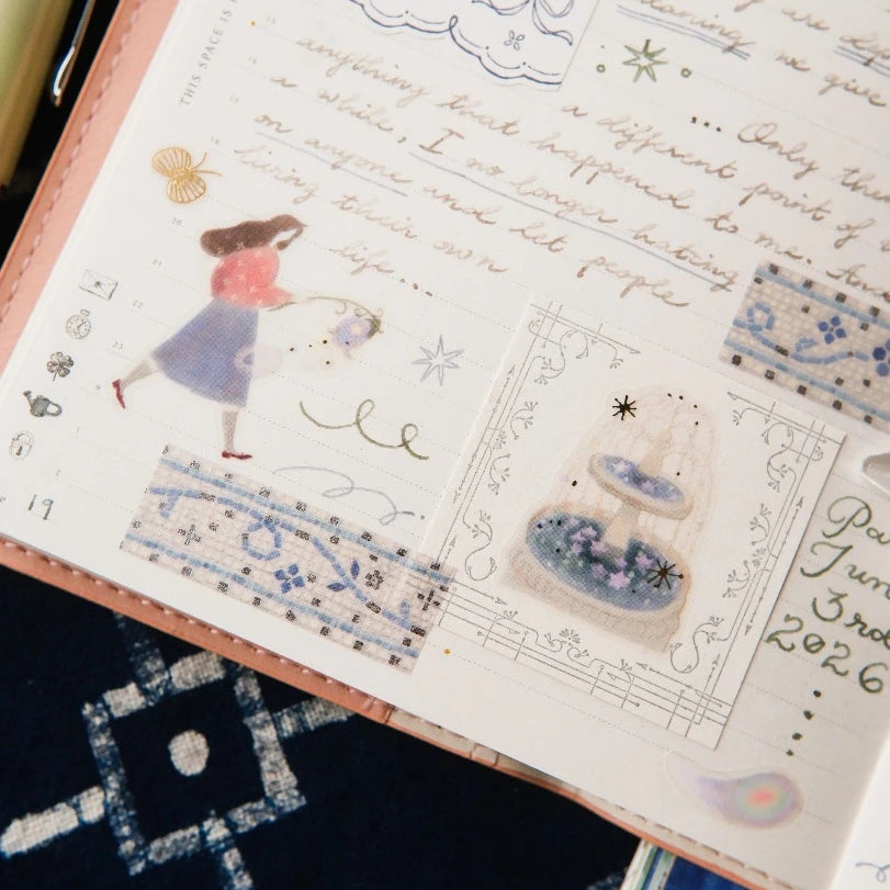 A close-up of a decorative planner or journal page with pastel blue and pink illustrated stickers, handwritten script, and patterned washi tape on a peach-colored notebook cover, resting on a dark patterned fabric surface.
