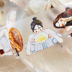 A close-up of a transparent tape roll featuring cute anime-style illustrations of women, including one holding a bowl of noodles and another holding a folding fan, with a soft-focus background.