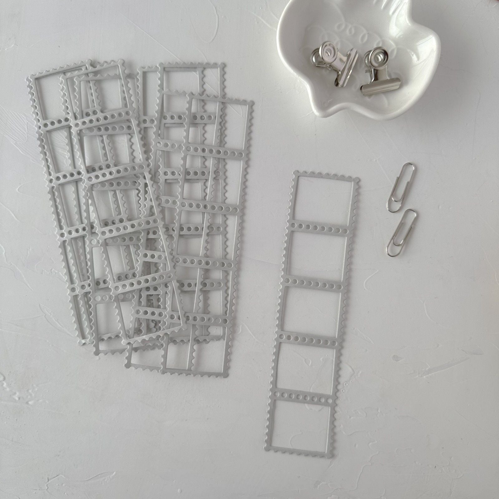Metal dies shaped like film strips on a light gray surface with a small container of clips.