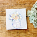 Watercolor painting of flowers in a vase on a wooden surface with real hydrangeas.