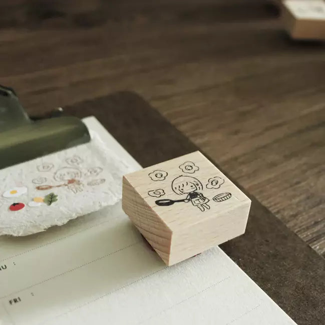 Mini Girls Frying Eggs stamp with its cartoon cooking design on a warm wooden surface