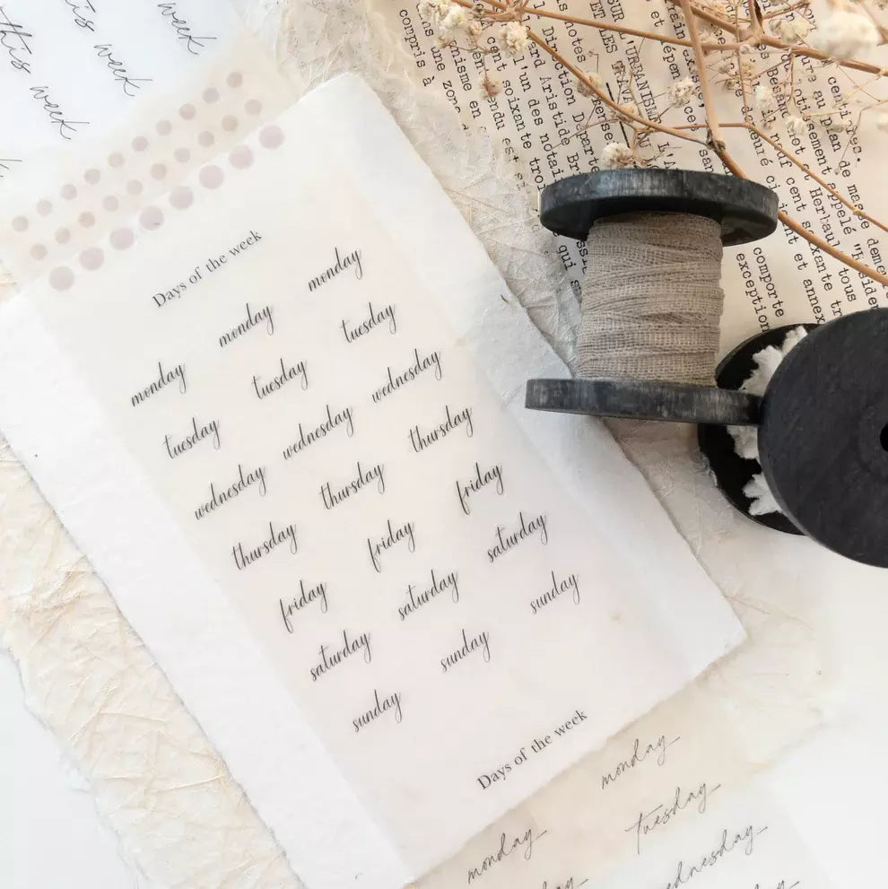 Sheet of paper with handwritten days of the week on a textured surface with a spool of thread.