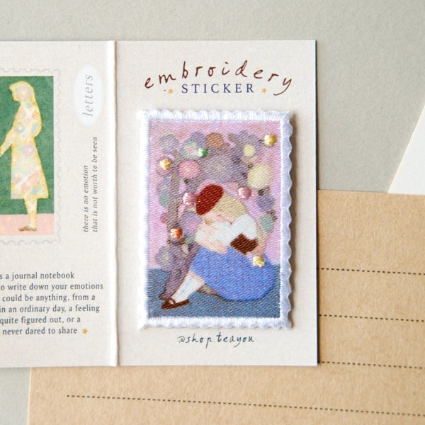 An embroidered sticker card featuring a stitched illustration of a girl with balloons, labeled 'embroidery STICKER' and signed 'shop tea time', displayed on a neutral surface next to a notebook page.