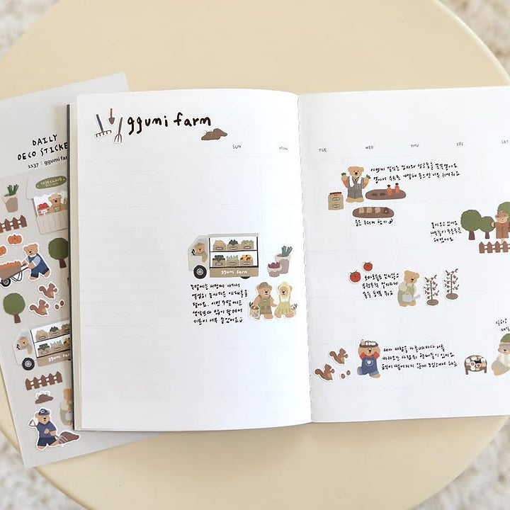 Open book with illustrated 'ggumi farm' pages on a beige surface, from the Ggumi Farm Sticker Sheet