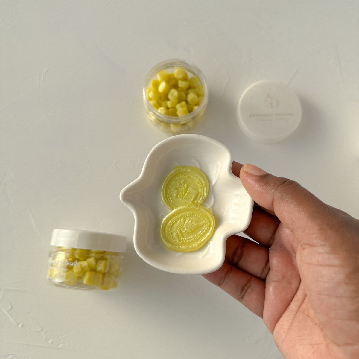 Hand holding a white dish of Golden Apple wax seal discs surrounded by small jars on a grey backdrop