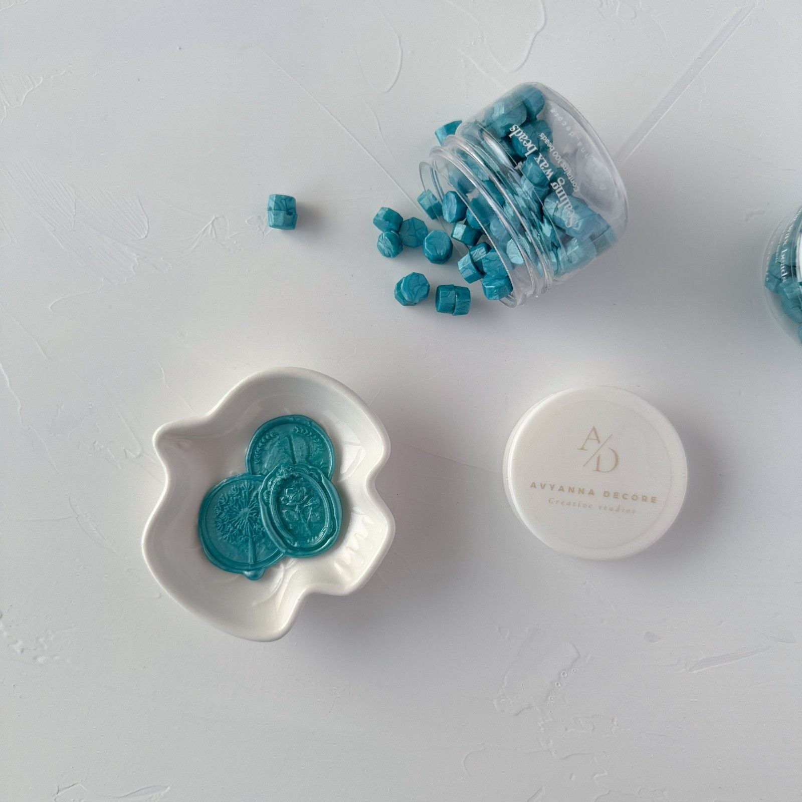 Teal decorative stones in a clear container and white dish on a light gray surface.