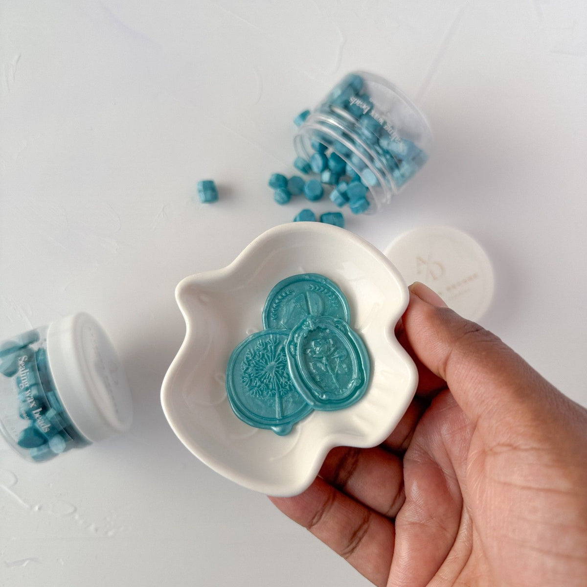 Hand holding a shell-shaped white dish of tropical Hawaiian Blue sealing wax beads on a grey backdrop