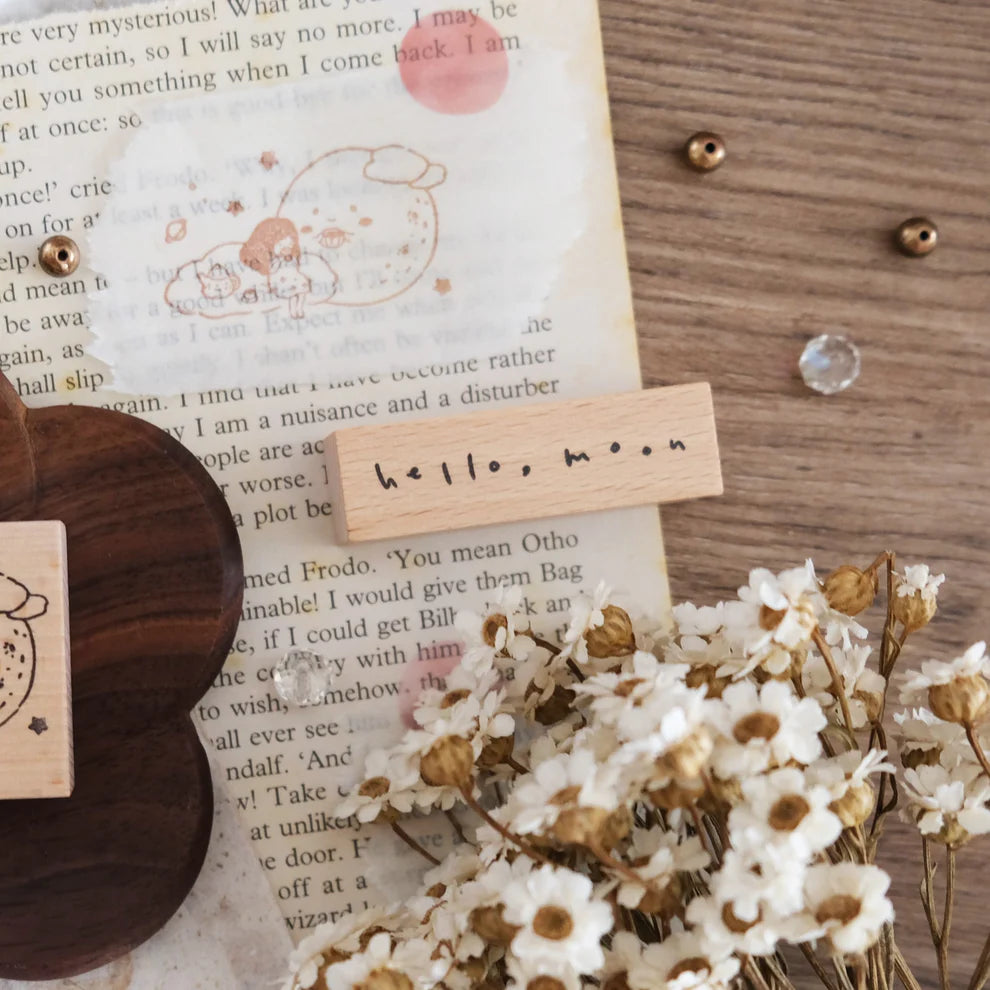 Hello Moon Rubber Stamp print on a wooden card resting on a book with dried flowers and stamps