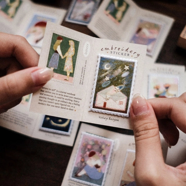 A person holds a small printed card featuring embroidered-style stickers with vintage illustrations, including a house under a full moon and a woman in a dress. Other similar sticker cards are scattered in the background on a dark surface.