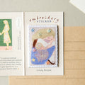 An open notebook or journal page with a small embroidered sticker centered on the right side. The sticker shows a pastel illustration of a woman and child with stars and floral accents.