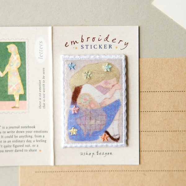 An open notebook or journal page with a small embroidered sticker centered on the right side. The sticker shows a pastel illustration of a woman and child with stars and floral accents.