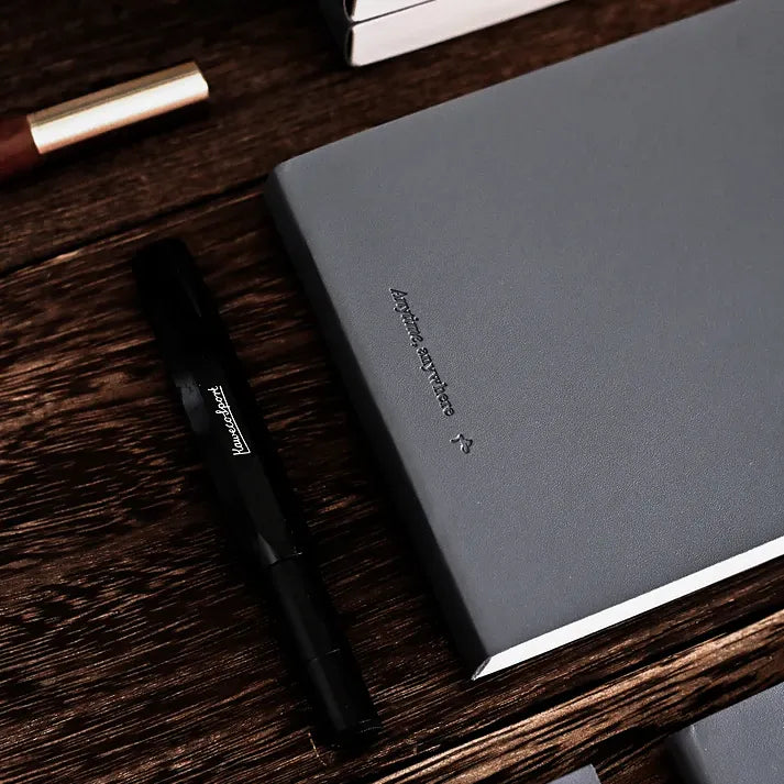 Dark grey Anytime Anywhere Journal closed with a pen resting on the cover, styled on a warm wooden desk