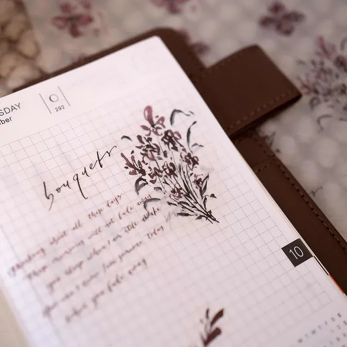Notebook styled with Floral PET Tape blooms and text on a floral-patterned backdrop, styled flat lay