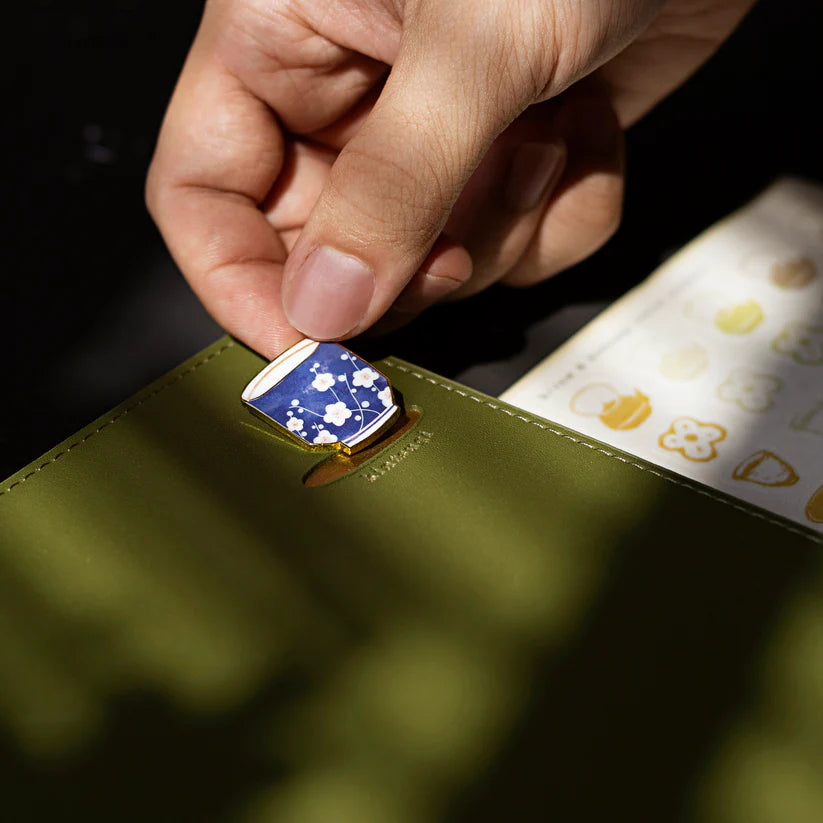 Hand holding a green leather product with a blue and white floral emblem, blurred background