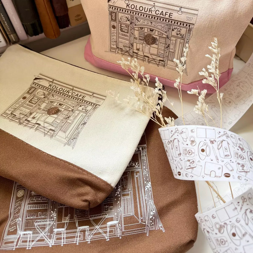 Textile items with 'Kolour Cafe' design on a surface with dried plants.