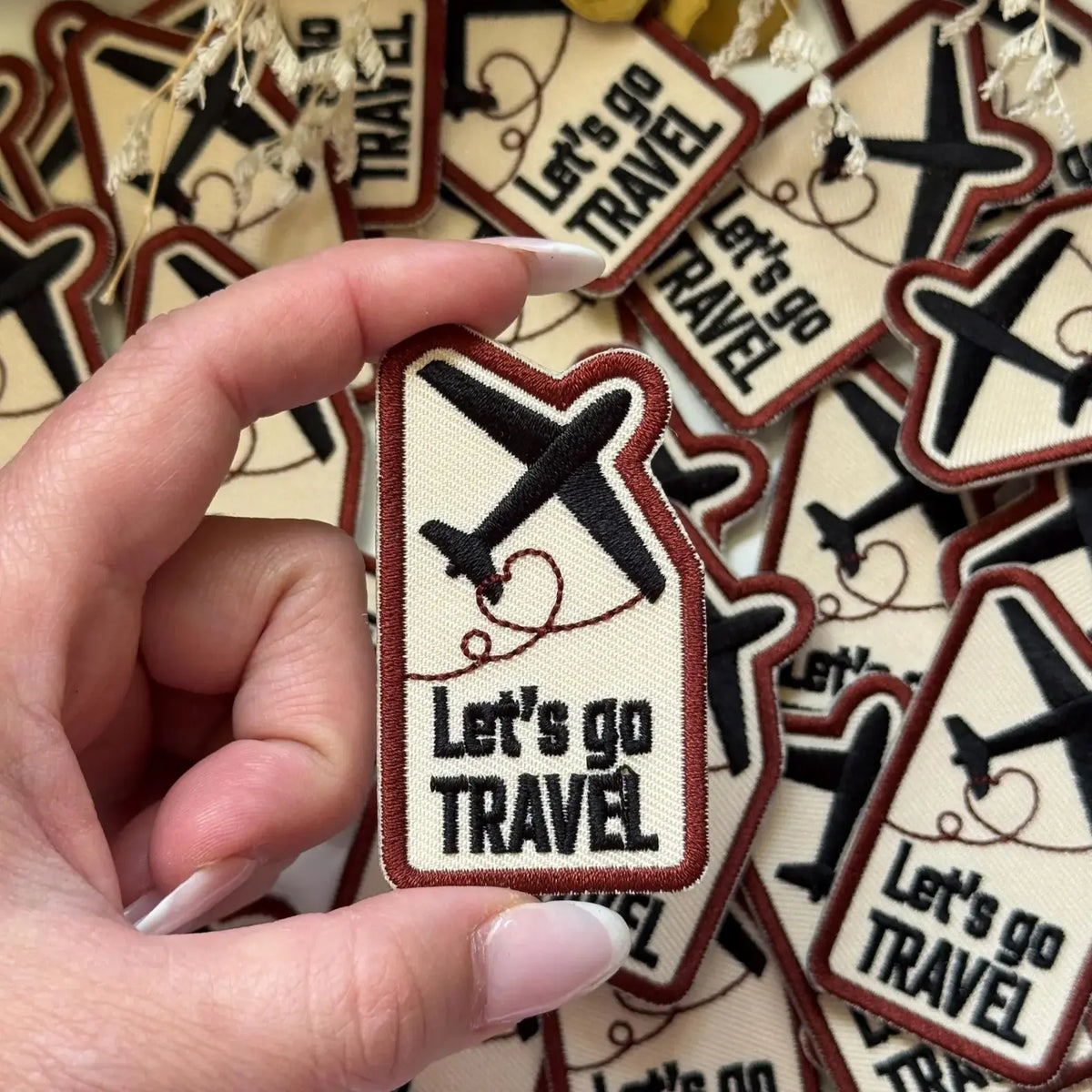 Hand holding a single Let's Go Travel Embroidered Patch with a stack in the background