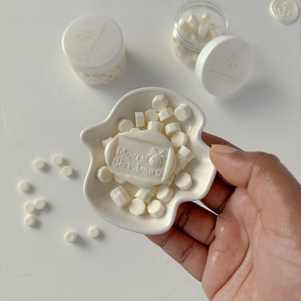 Hand holding a Merry Christmas stamped Light Ivory wax seal with creamy cube beads on a light backdrop