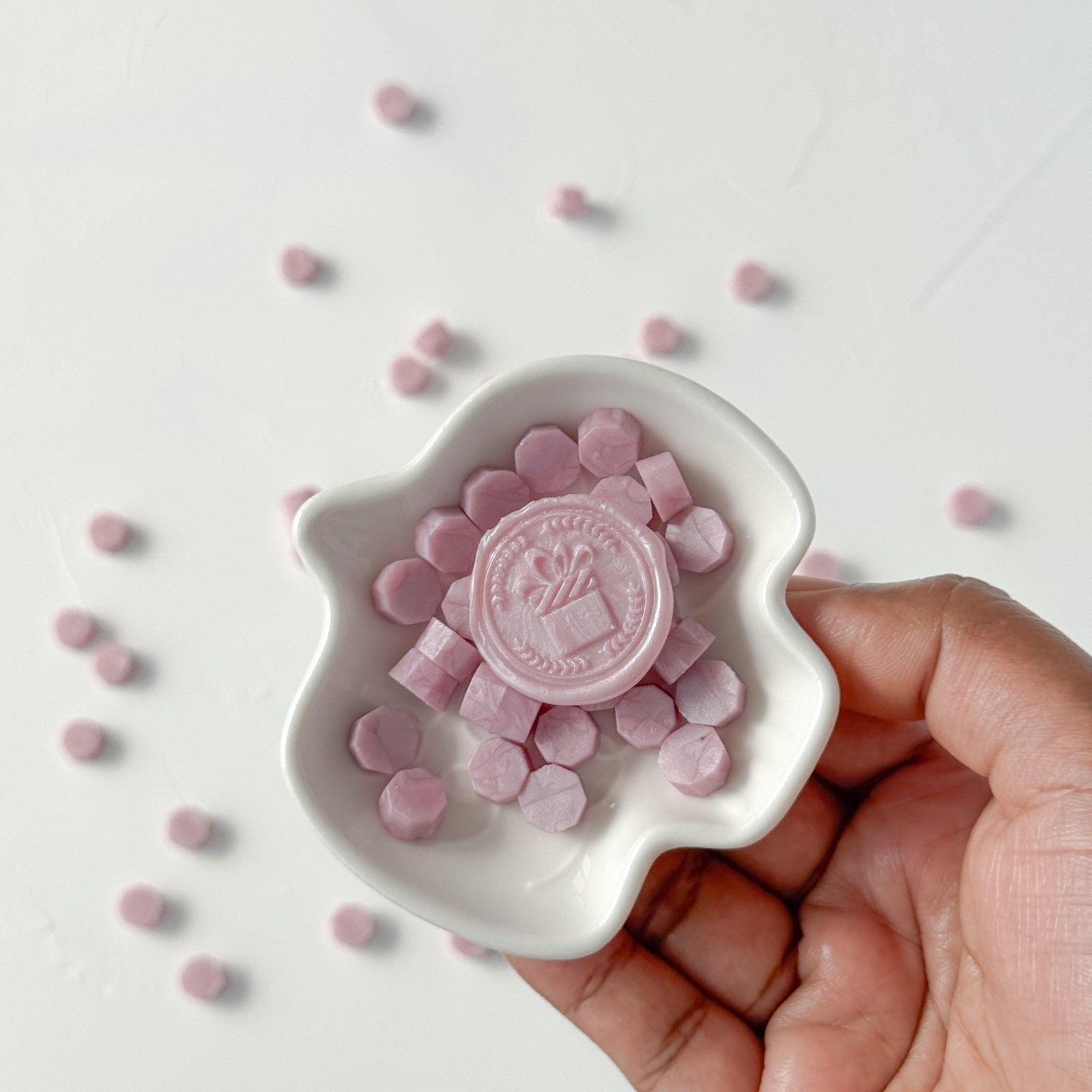 Hand holding a white dish with pink candy and a visible brand logo on a light background