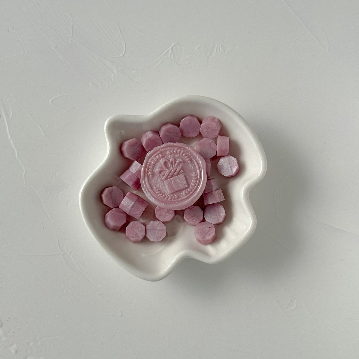 White dish of pastel Light Lavender Wax Beads with a stamped logo seal on a light grey studio background