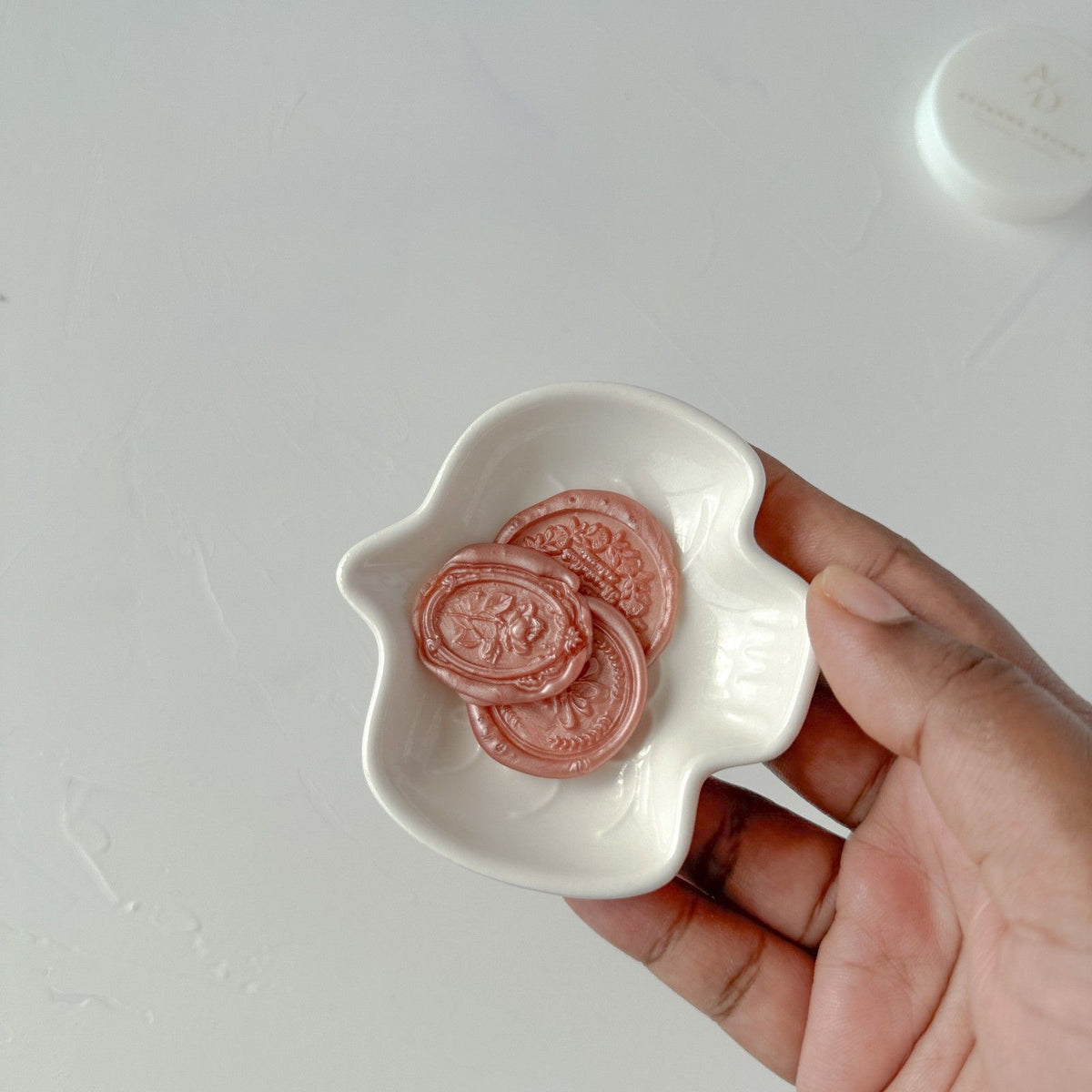 Hand holding a small white dish filled with round Lingering Brew Wax Beads against a light grey background