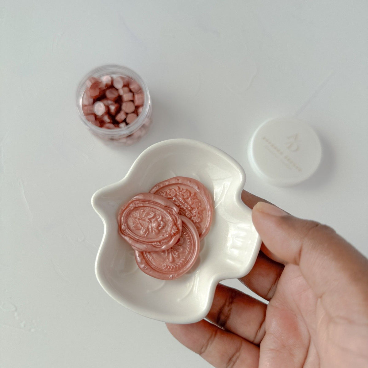 Hand presenting a dish of Lingering Brew Wax Beads with loose beads scattered around on a light grey surface