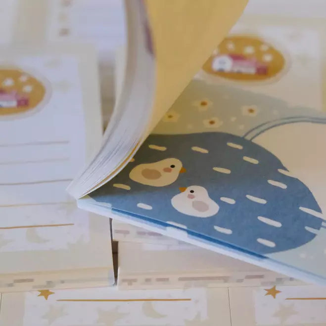 Little Village Memo Pad open to show its cartoon character pages on a wooden surface