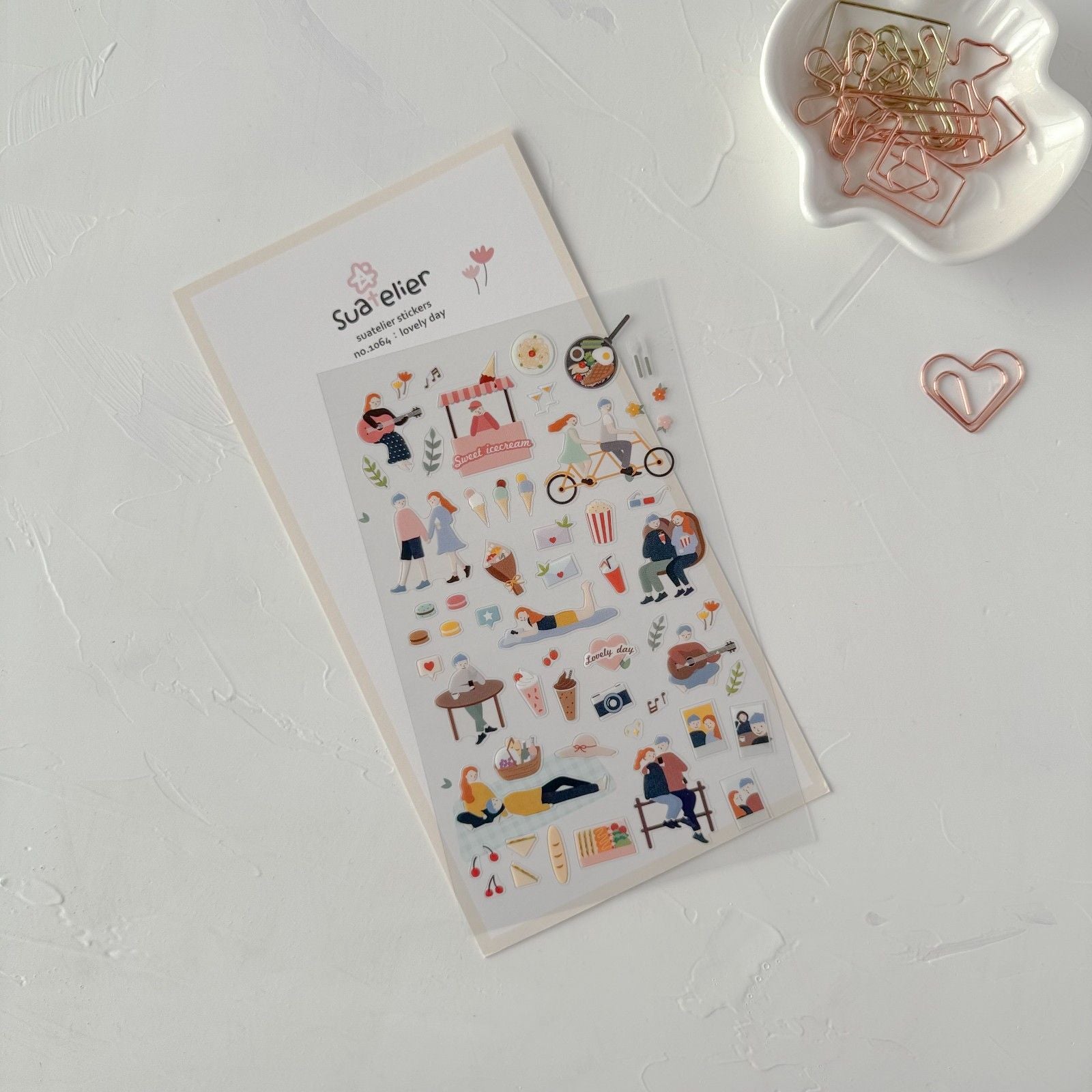 Sheet of decorative stickers with illustrations on a white surface, next to a small container of paper clips.