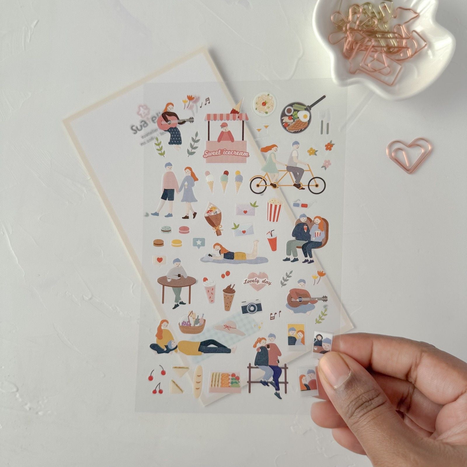 Sheet of illustrated stickers held by a hand on a light surface with decorative items.