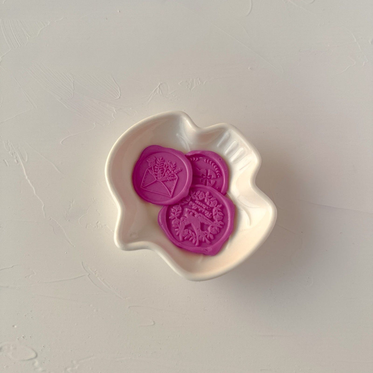 Three stamped Magenta Pink wax seals with a decorative design in a white dish on a light grey backdrop