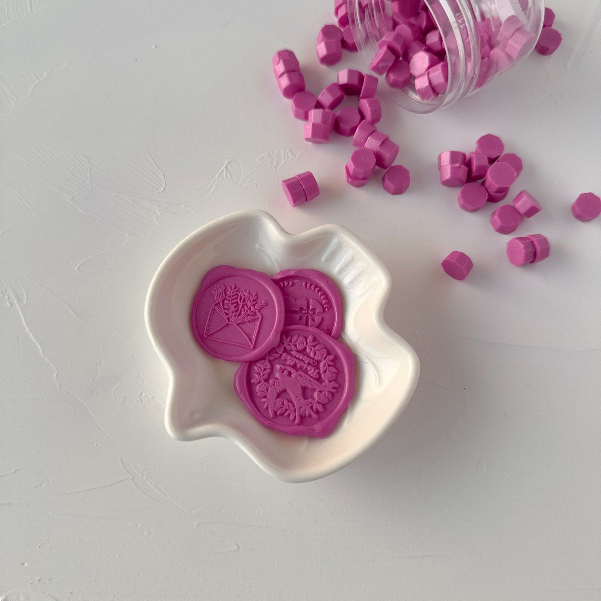 Hexagonal Magenta Pink sealing wax beads piled in a white dish on a light grey background, top view