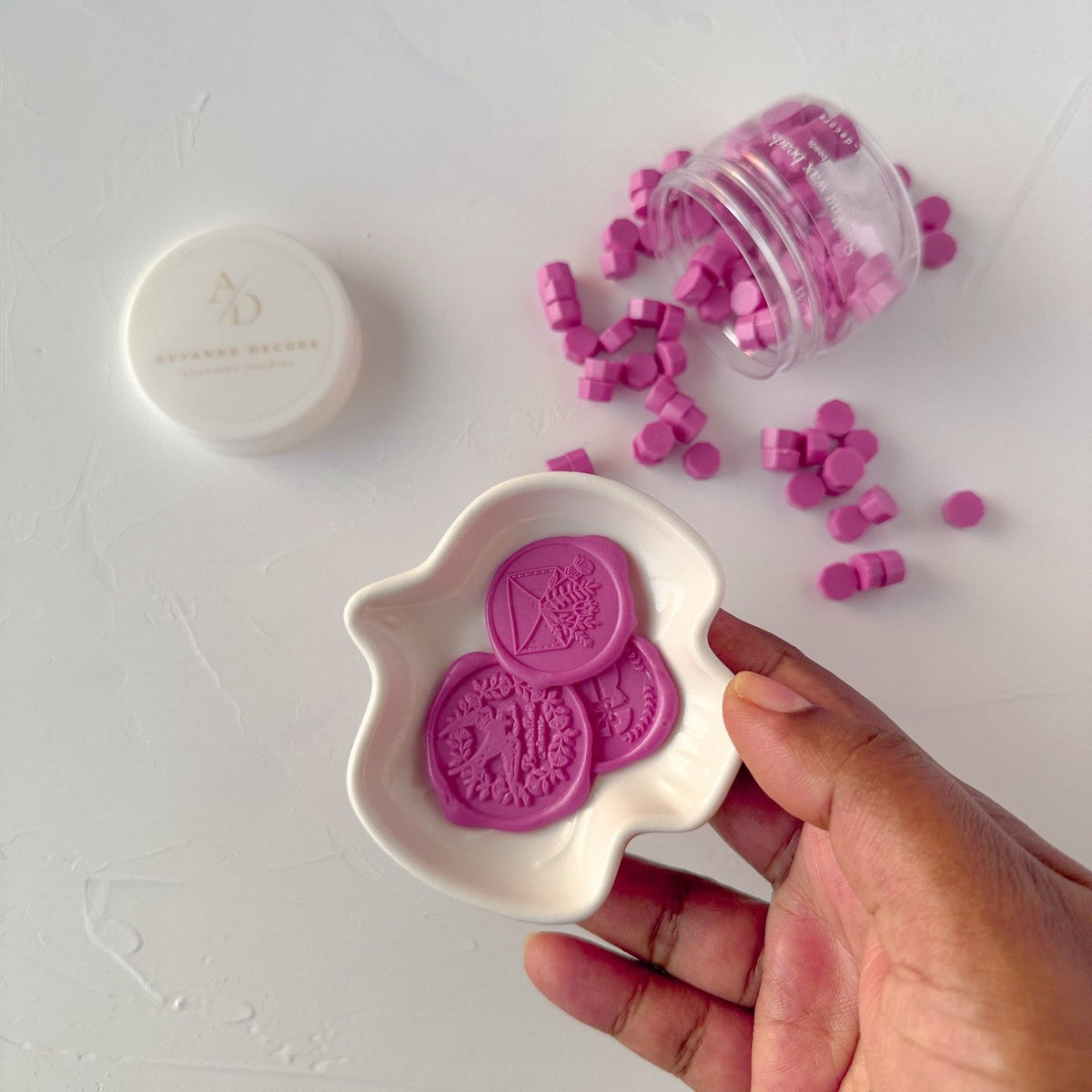 Hand holding a white mold of Magenta Pink Wax Beads with loose bead pieces around on a grey backdrop