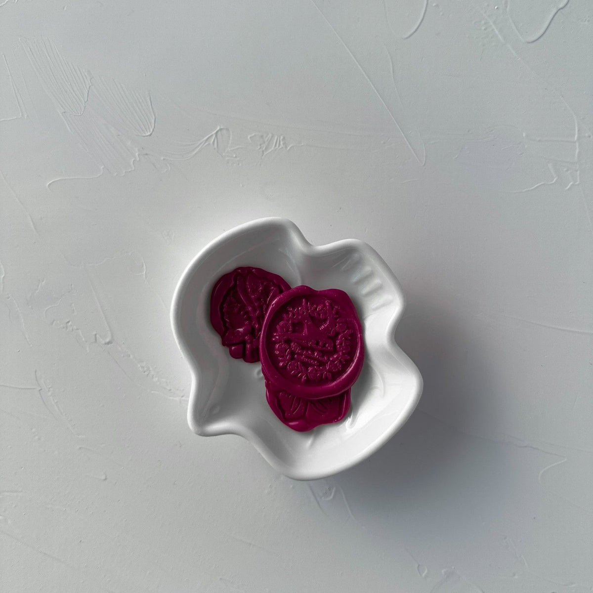 Two stamped Magenta Rush wax seals displayed in a white bowl on a soft grey background, top-down view