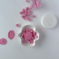 Pink wax seals on a white dish with a container of additional seals on a light gray background.