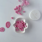 Pink wax seals on a white dish with a container of additional seals on a light gray background.
