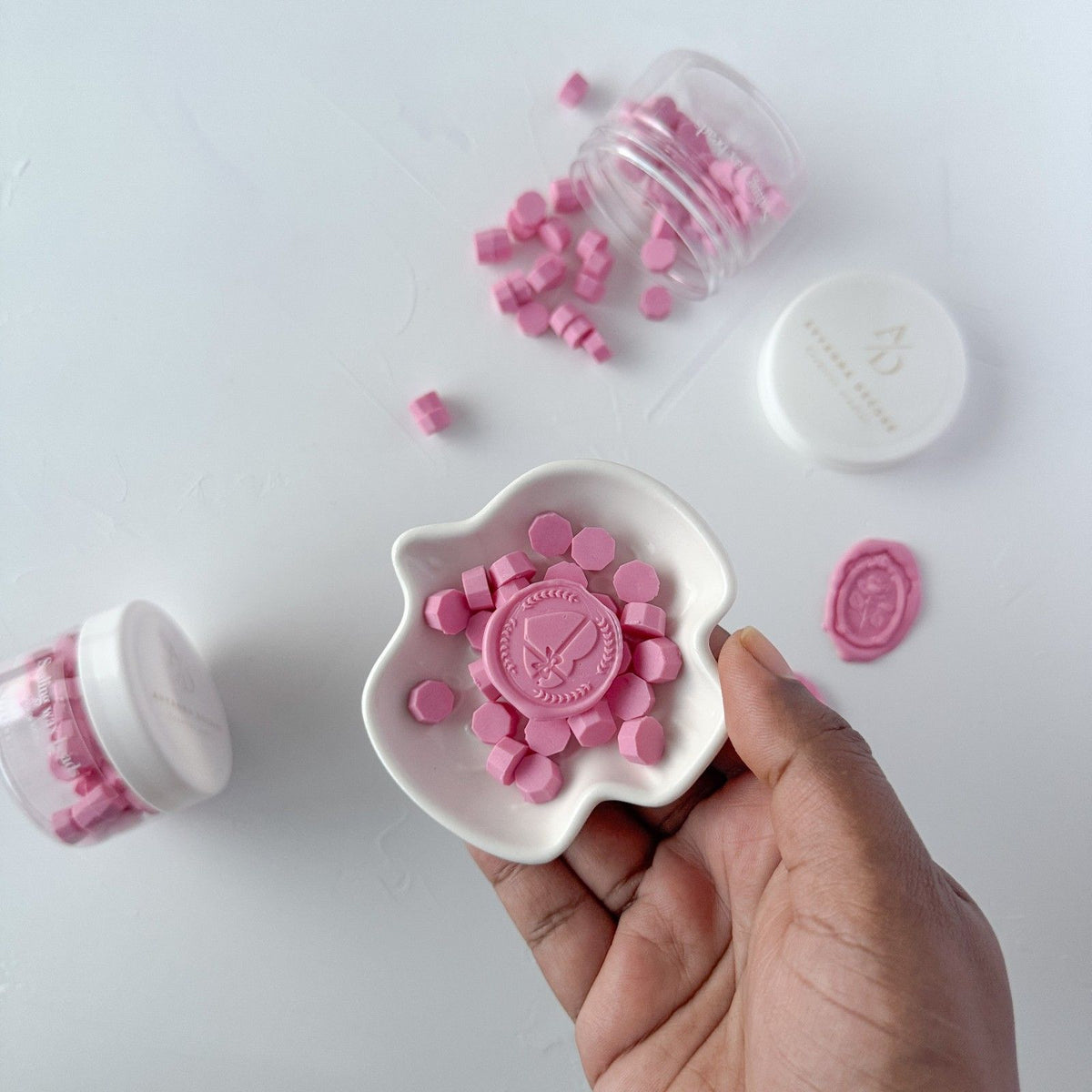 Hand holding a white container of stamped Maiden Pink wax seals against a light grey studio background