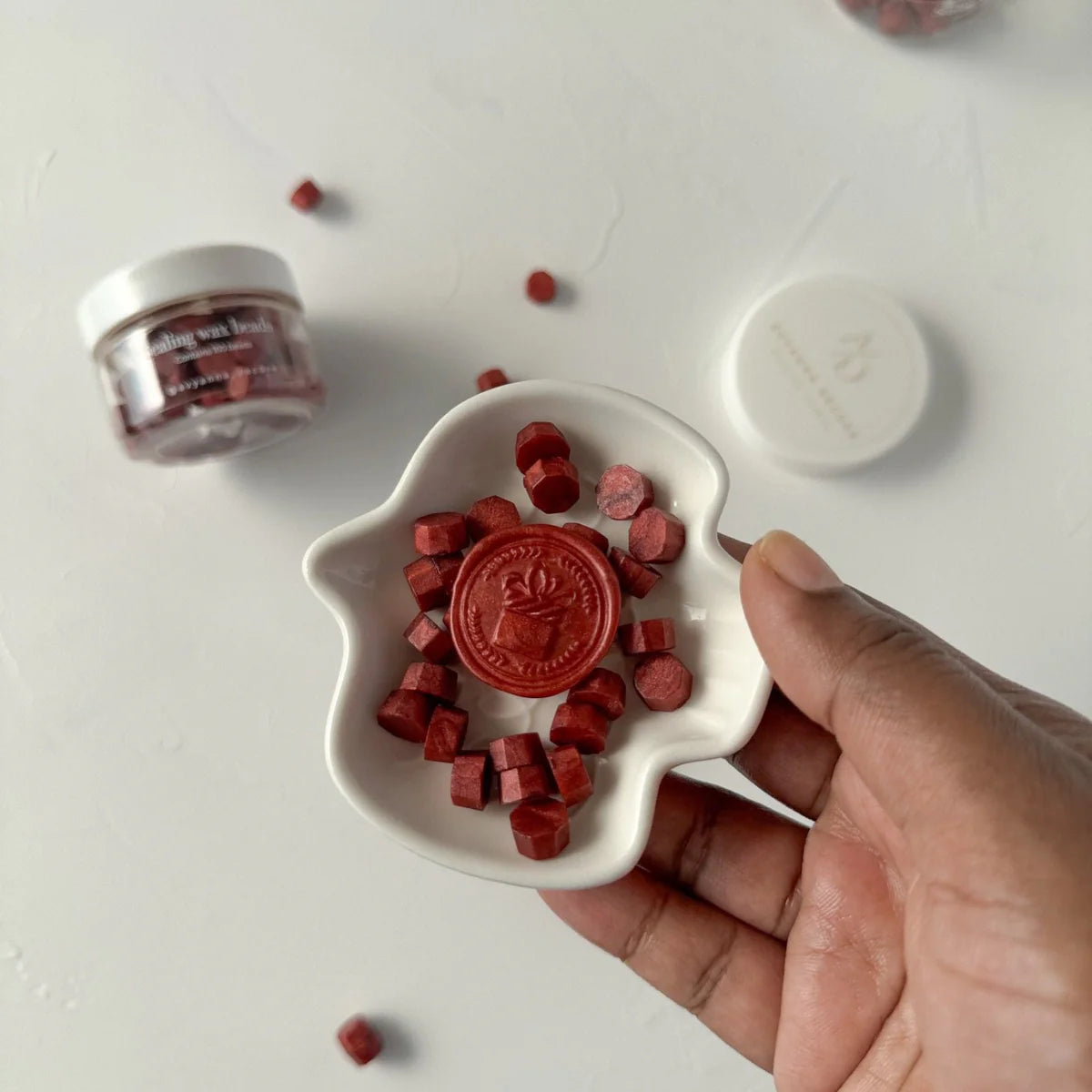 Hand holding a dish of Maroon Wax Beads cubes with the storage jar and lid behind on a white backdrop