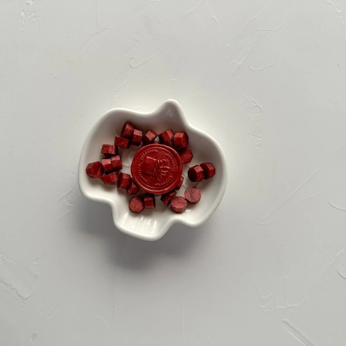 Small white dish of hexagonal deep maroon sealing wax beads on a light grey studio background, top view