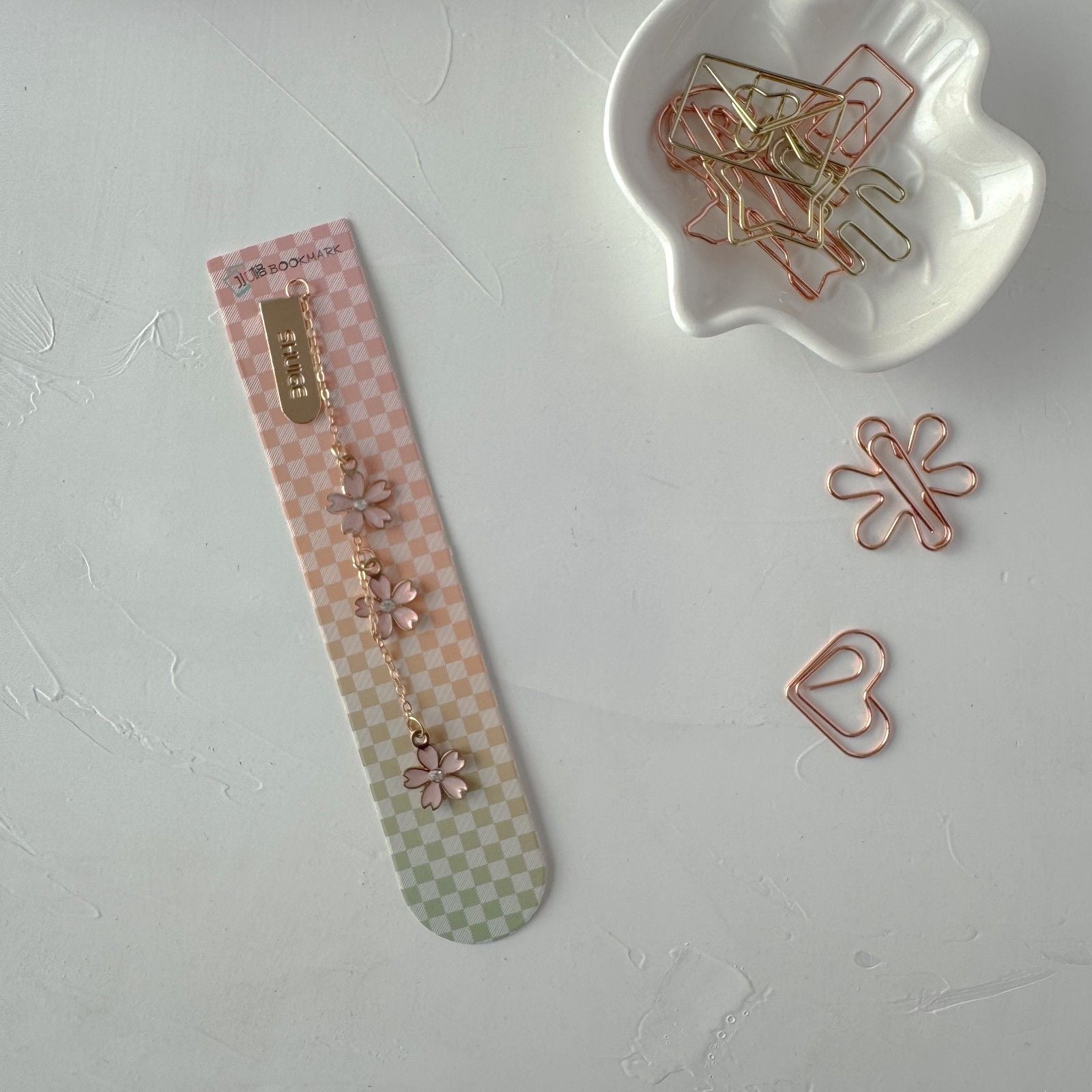Decorative paper clips on a checkered strip with a white dish containing more paper clips on a light gray background.