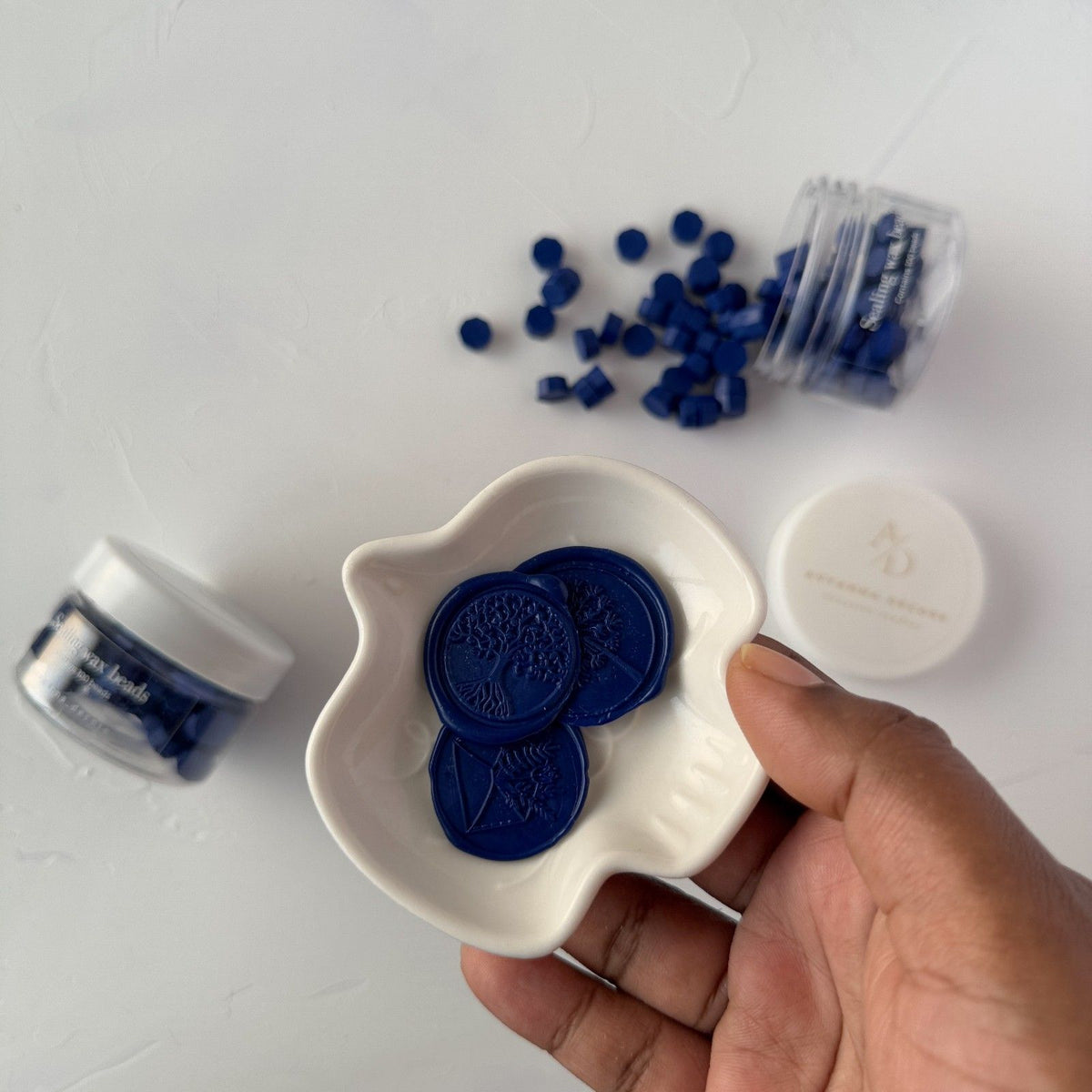 Hand holding a white dish of deep navy Midnight Blue sealing wax beads on a light grey background