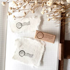 Wooden stamp with postal symbol on a piece of paper with similar stamp, surrounded by dried flowers.