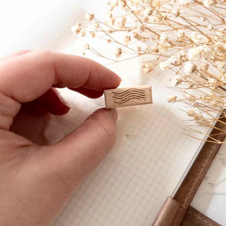 Hand holding the small wooden Mini Postal II Rubber Stamp against a floral background
