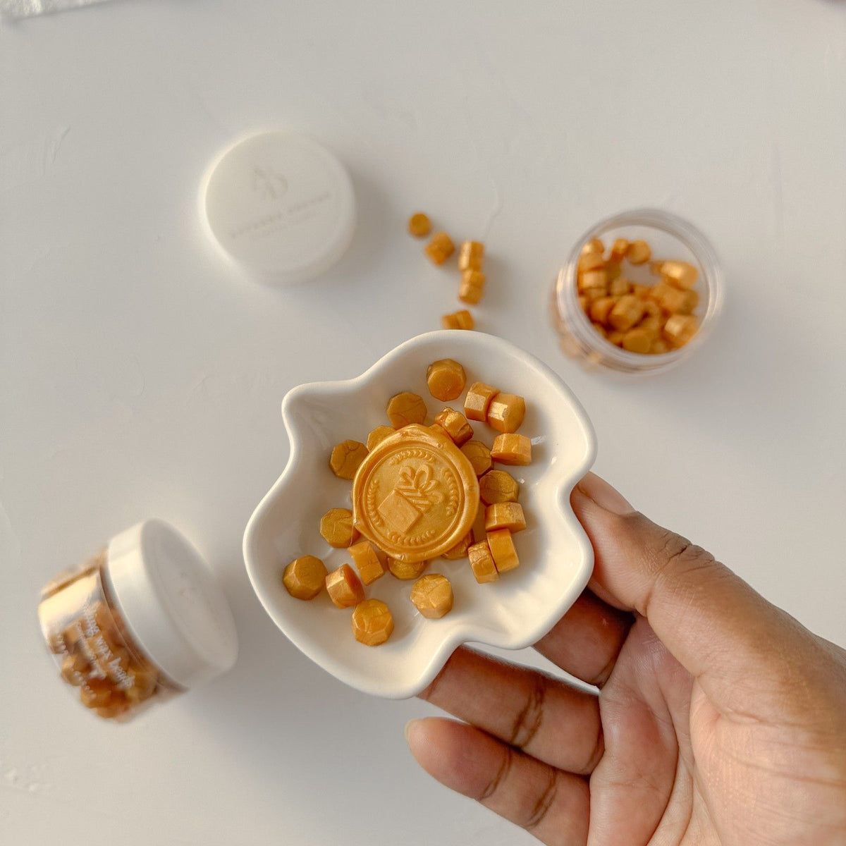 Hand holding a dish of Mustard Orange Wax Beads with loose beads scattered around on a light background