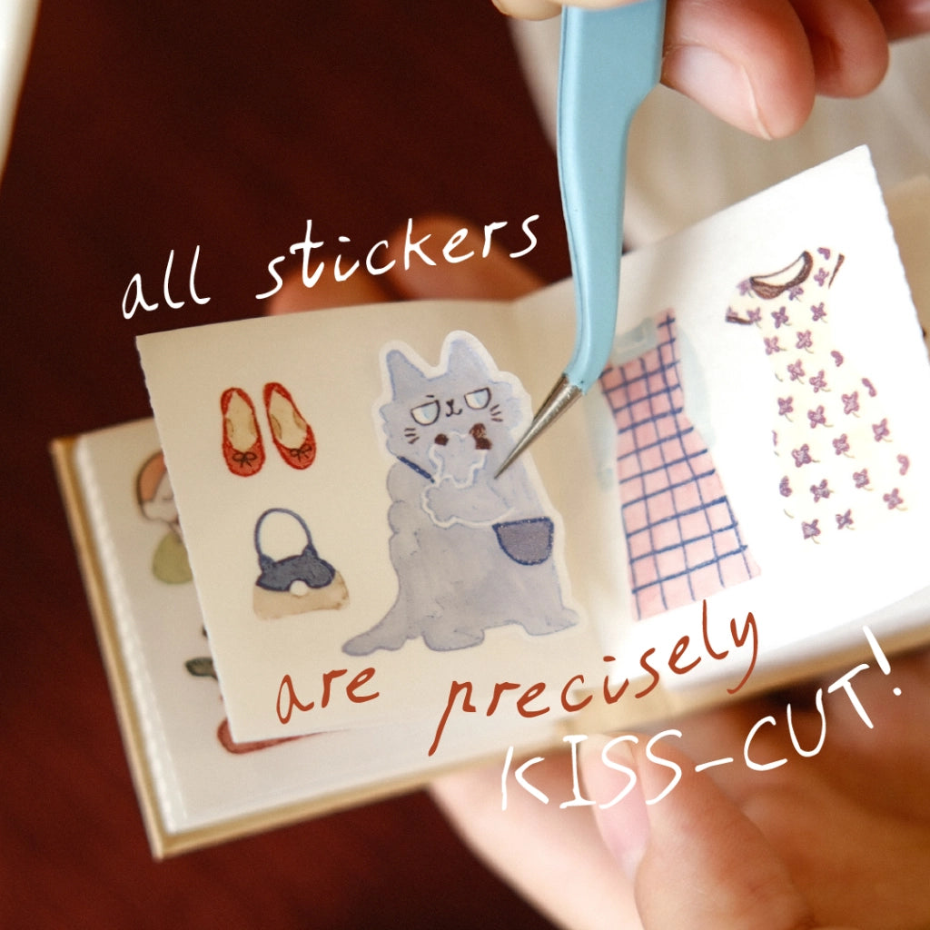 Sticker book with various stickers, including a cat, shoes, and plaid patterns, held by a hand with a cutting tool.