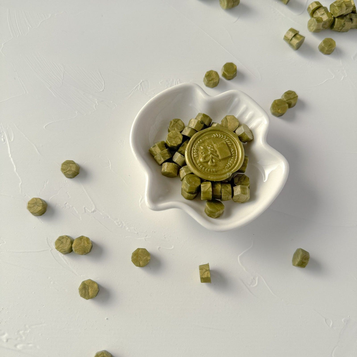 Shell-shaped white dish of muted green Olive Wax Beads with loose beads scattered on a white background