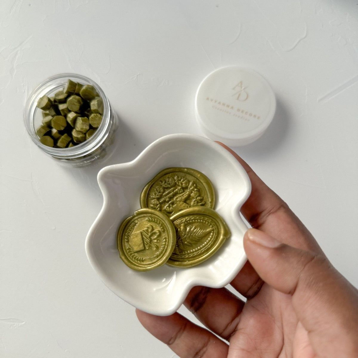 Hand holding a dish of Olive Wax Beads seals with the jar and white container on a light background