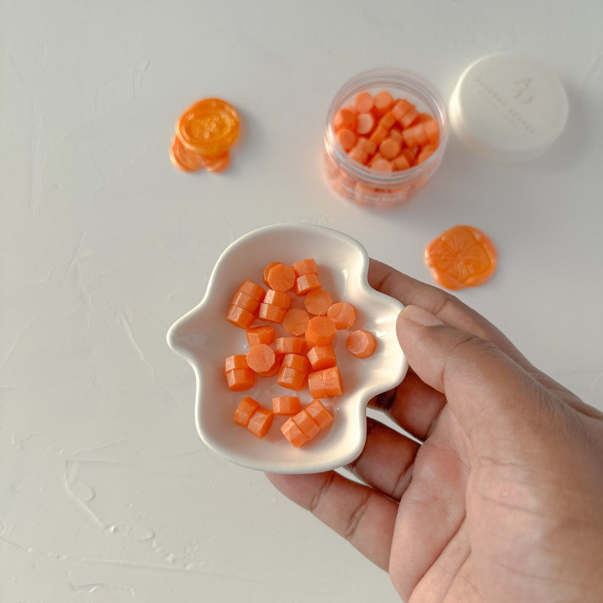 Hand holding a white leaf-shaped dish of carrot-shaped Orange sealing wax beads on a grey background