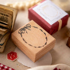 Wooden rubber stamp with a hand-drawn floral wreath design, placed on a craft table beside wax seals and vintage-style paper packaging.