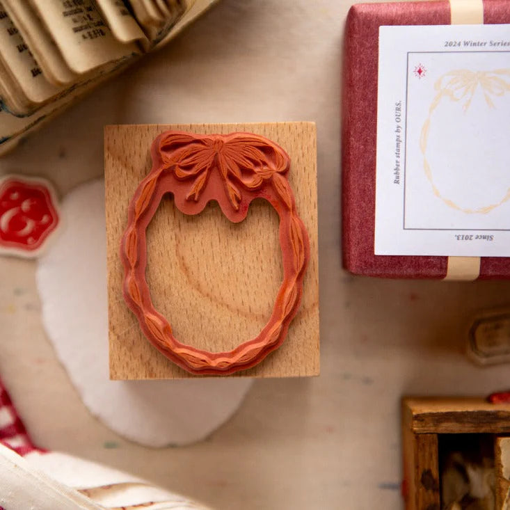 Carved rubber stamp with a bow-topped wreath design mounted on a wooden block, displayed on a craft workspace with wrapped stationery items, wax seal accents, and vintage paper elements.